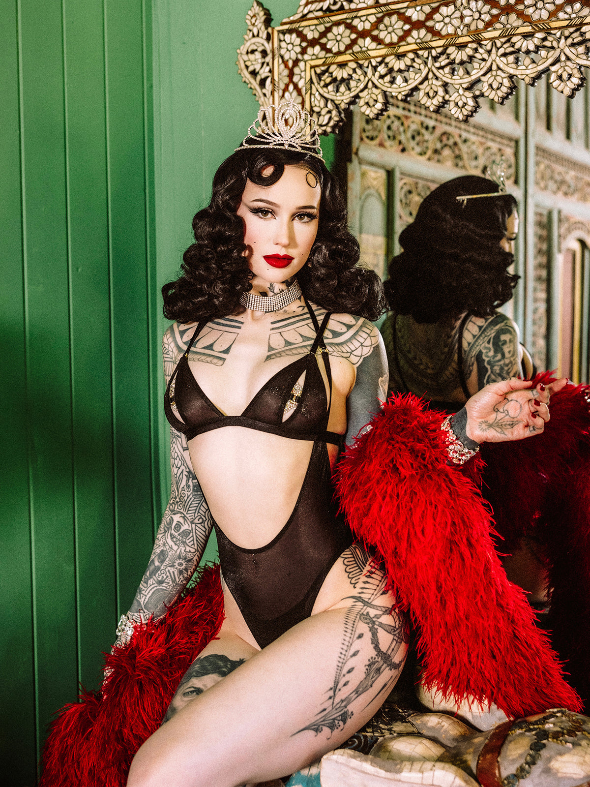 A woman with dark curly hair, tattoos, and a tiara poses in Frederick's of Hollywood's Pia Shine Mesh Open Cup, Backless Teddy & Bralette Set, styled with a red feathered robe, against a decorative mirror and green wall.
