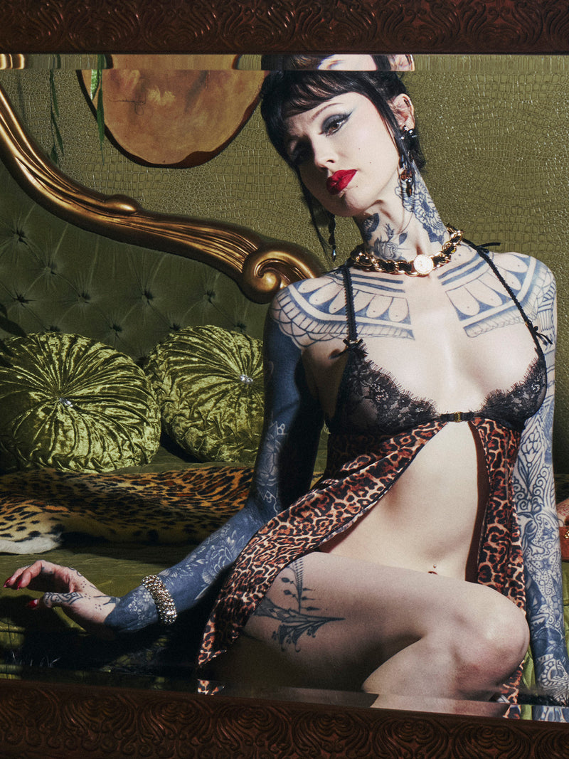 A heavily tattooed woman models the Frederick’s of Hollywood Raven Leopard And Lace Babydoll Set, featuring a black lace bralette and leopard print robe, while posing on a green velvet tufted sofa with gold frame. She accessorizes with red lipstick and a gold chain necklace.