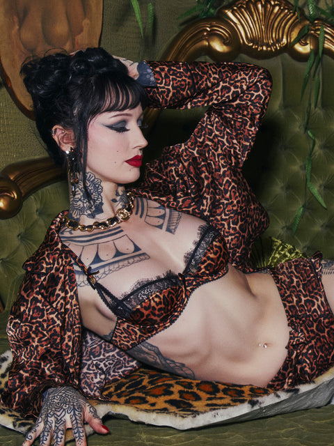 A person with dark hair and extensive tattoos reclines on an olive green tufted sofa, posing against a moody, vintage backdrop. They wear the Frederick’s of Hollywood Raven Leopard And Lace Bra with matching leopard-print robe and bottoms, accented by a gold chain necklace and bold makeup.