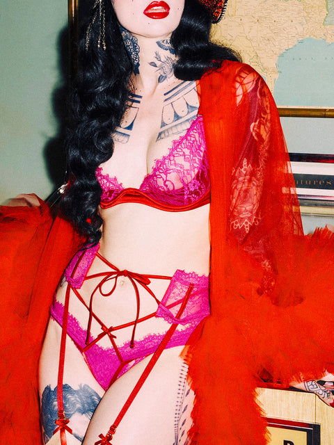 A person with long black hair and tattoos models the Rosalind Garter by Frederick's of Hollywood—bright pink lace lingerie with red straps and a sheer red robe—in front of map and art, face partly seen with bold red lipstick.