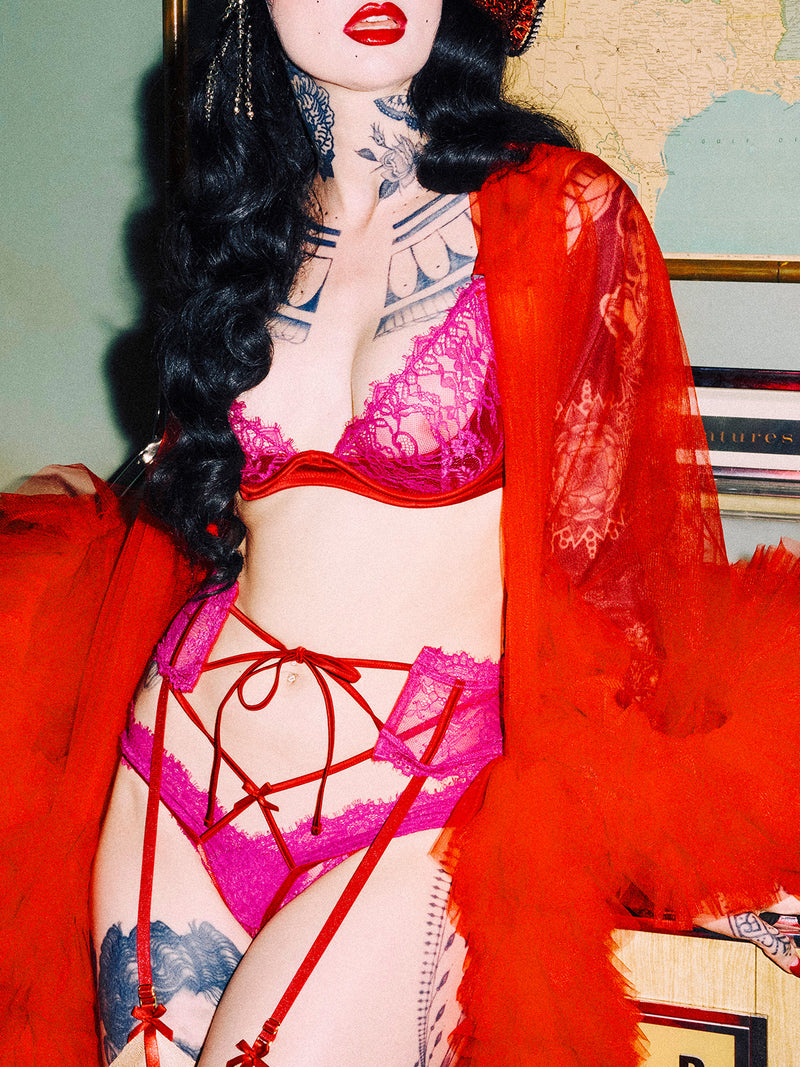 A person with long black hair and tattoos models the Rosalind Garter by Frederick's of Hollywood—bright pink lace lingerie with red straps and a sheer red robe—in front of map and art, face partly seen with bold red lipstick.