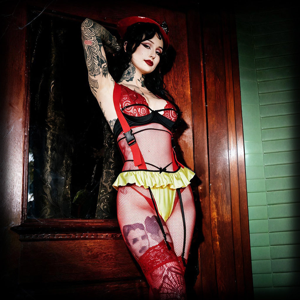 A tattooed woman poses indoors wearing a revealing red and black lingerie set with a bright yellow ruffled underskirt and red fishnet stockings.