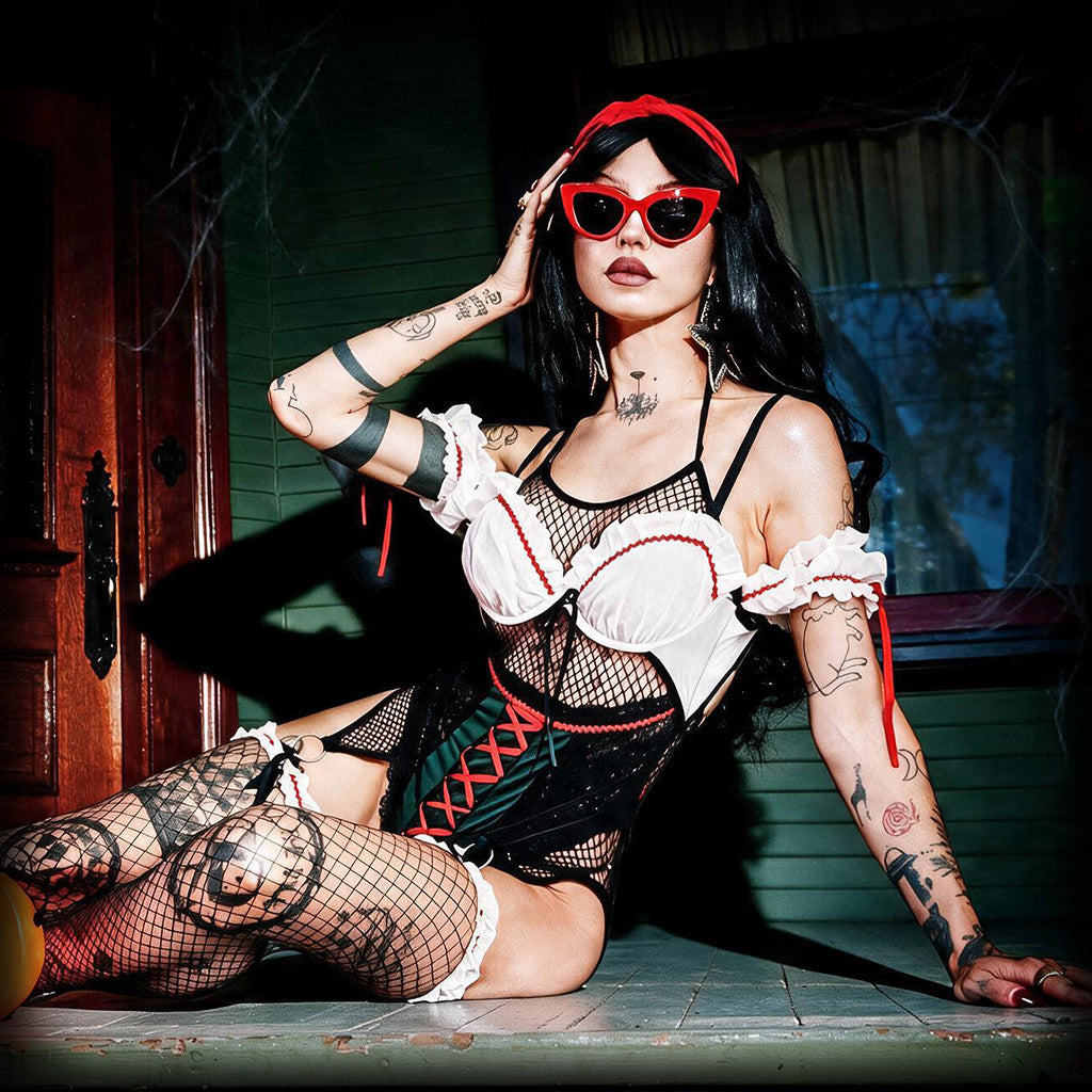 A tattooed woman wearing a gothic-style lingerie outfit with a white ruffled corset top, black fishnet bodysuit, red trim, and red heart-shaped sunglasses, posing on a porch floor.