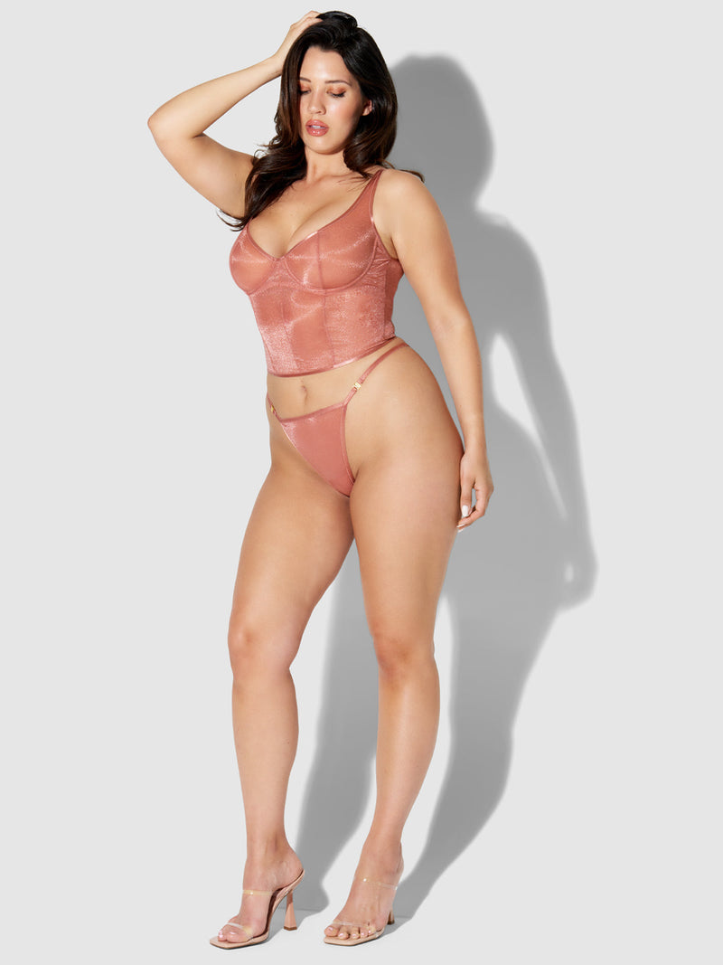 A woman poses against a light gray background wearing the Frederick's of Hollywood Pia Mesh G-String in dusty pink with an embossed waistband and clear high heels, one hand touching her head, effortlessly showcasing sheer elegance.