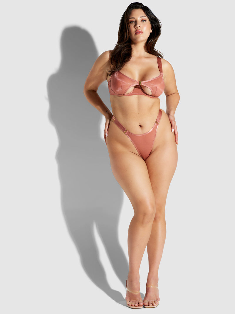 A woman poses confidently in a blush pink bikini and clear heels against a light gray background, her look completed with the Pia Elastic Side Mesh Thong by Frederick's of Hollywood, adding sexy lingerie appeal as she casts a shadow to her left.