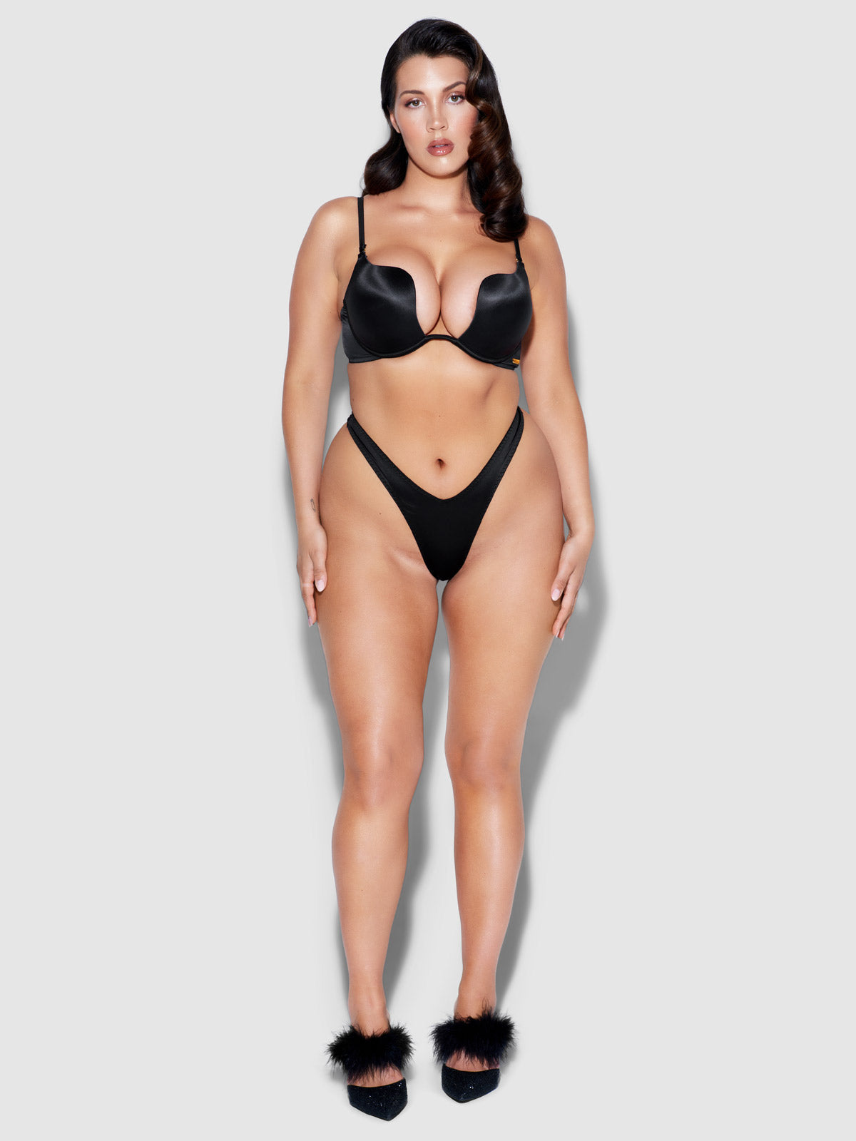A woman with long, dark wavy hair models Frederick's of Hollywood’s Rising Star Liquid Satin Tanga panties and a soft black satin bra, paired with black feathered heels, against a plain light gray background.