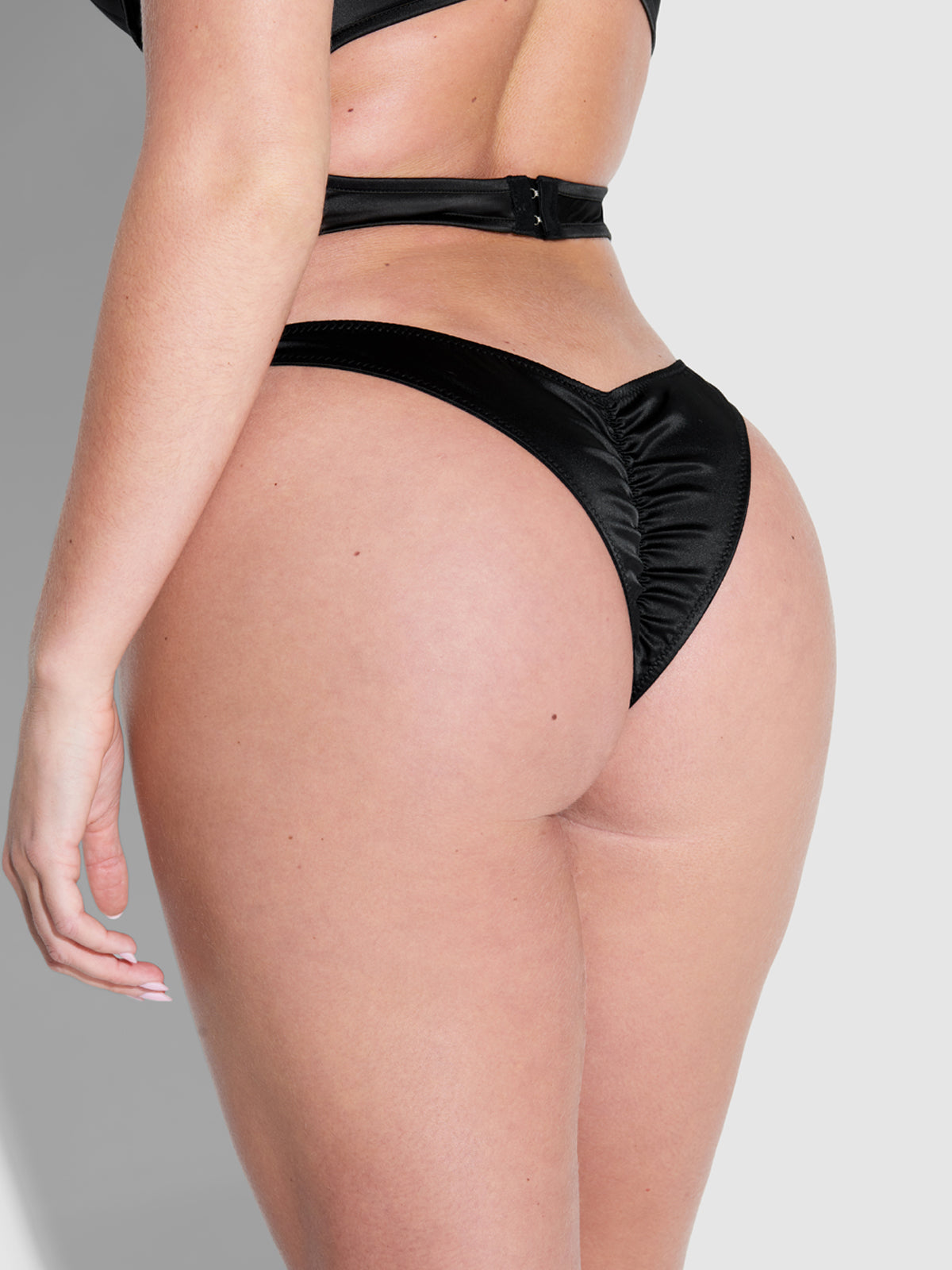 A model wears the Frederick's of Hollywood Petra Crotchless Satin Tanga, shown from the side and back against a plain light gray background, highlighting the lower back, hips, and upper thigh.