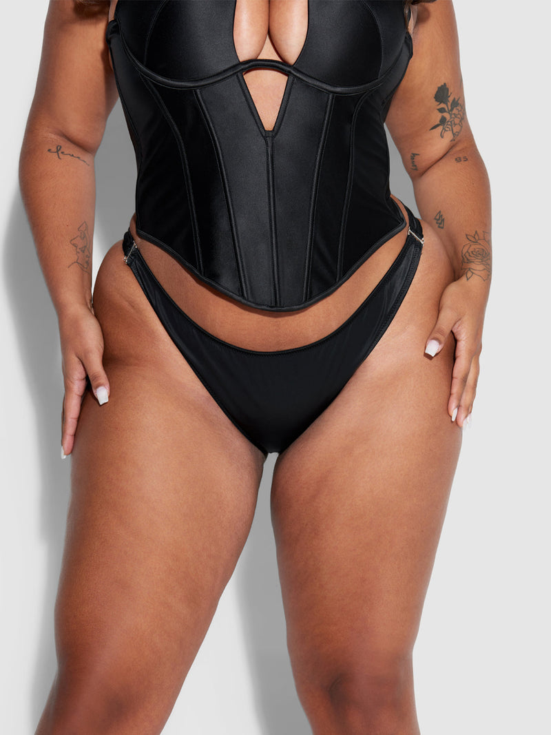 A woman models the Frederick's of Hollywood Petra Crotchless Satin Tanga, paired with a black corset-style top. Hands on hips, she displays tattoos on her arms, highlighting the lingerie’s silky finish and modern design.