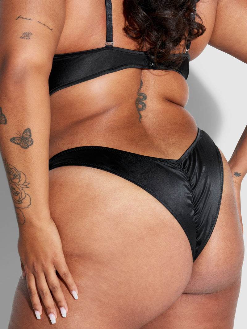 A close-up features a person wearing Frederick's of Hollywood’s Petra Crotchless Satin Tanga and black satin bra, highlighting their back and hip tattoos—a snake, butterflies, and flowers—as their hand rests gently on their hip.