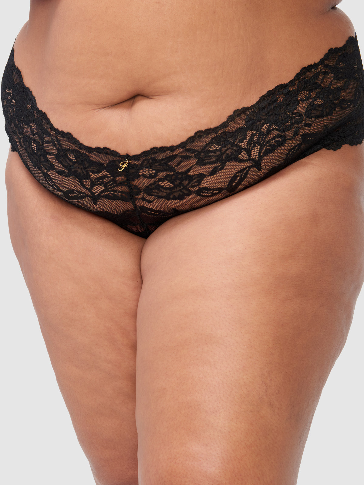 A close-up of a plus-size person wearing the Frederick's of Hollywood Mabel Crotchless Lace Up Back Cheeky, showcasing the black floral lace and gold detail at the waistband against a plain light background.