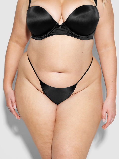 A plus-size woman models the Frederick's of Hollywood Lexi Liquid Satin G-String in black, shown from shoulders to mid-thigh against a plain background.