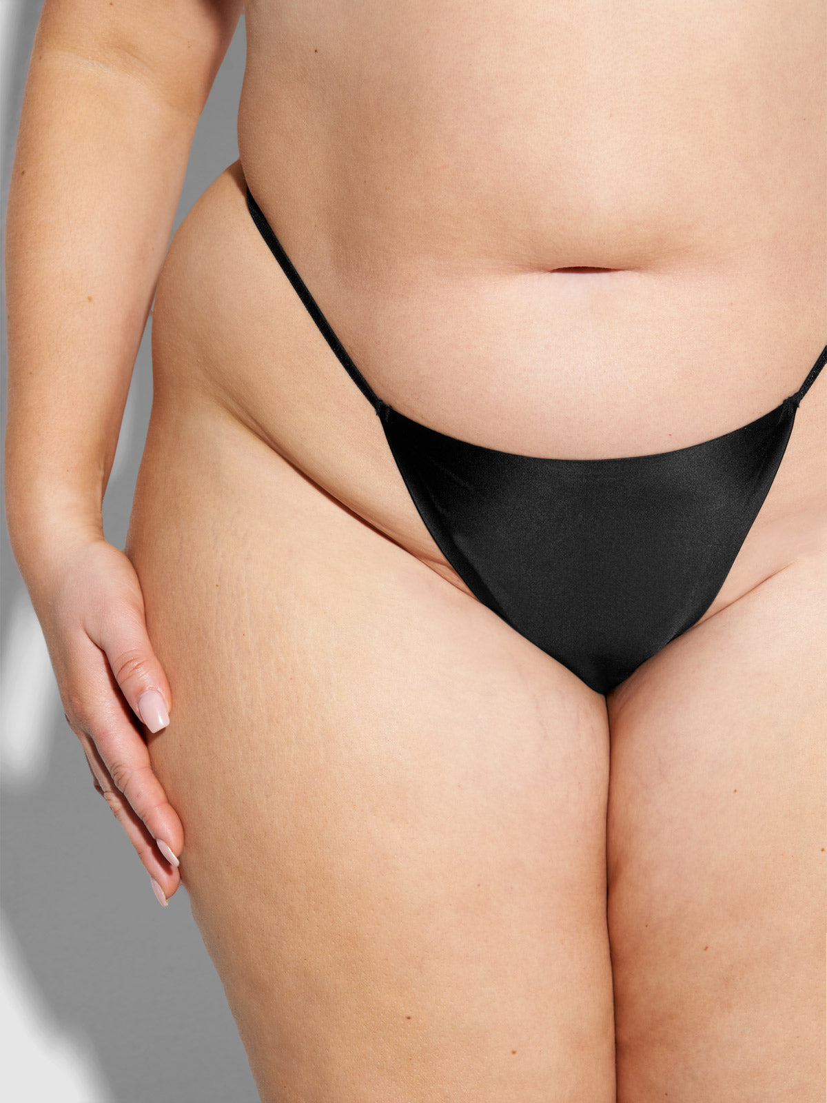 A close-up shows a person with light skin wearing the Frederick's of Hollywood Lexi Liquid Satin G-String, highlighting their midsection, hips, and upper thighs. Their hand rests on their thigh, emphasizing natural skin texture and stretch marks.