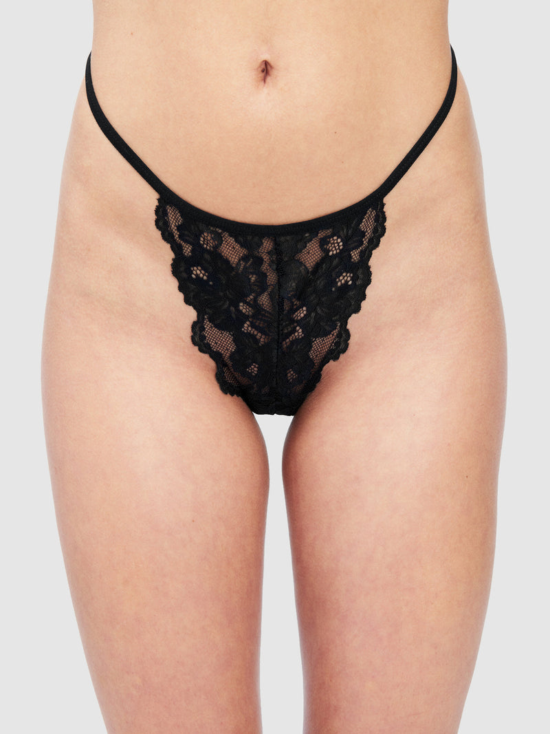 A person models the Mabel Lace G-String by Frederick's of Hollywood—a feminine black floral lace panty—shown from waist to upper thighs against a plain light background.