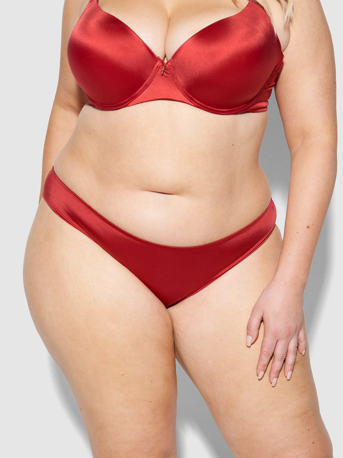 A woman poses against a plain light gray background, wearing the Frederick's of Hollywood Lexi Liquid Satin Hipster and matching bra, with one arm relaxed by her side.