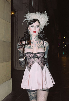 A person wearing a pink lace slip dress, patterned stockings, black lace gloves, and a feathered headpiece stands on a dimly lit street at night, holding sunglasses. They have dark wavy hair, bold makeup, and visible tattoos.