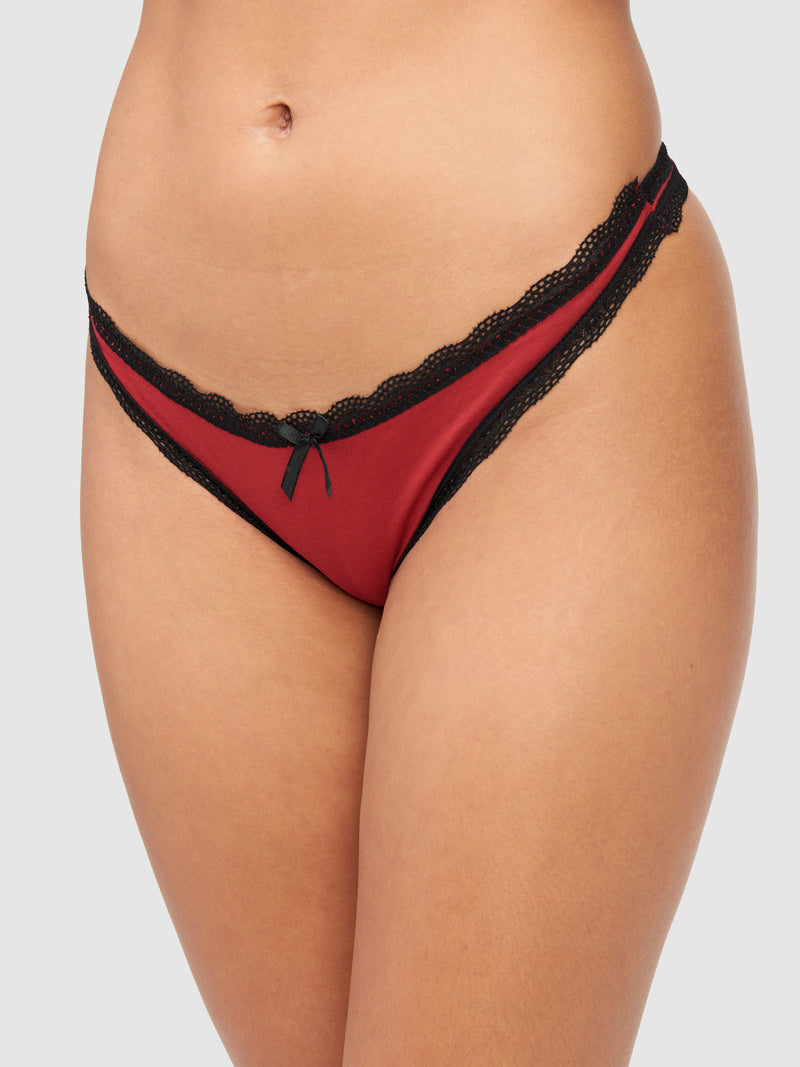 A close-up of a person wearing the Cora Cotton & Lace Thong by Frederick's of Hollywood in red, featuring black lace trim and a small black bow at the front, set against a plain, neutral background.